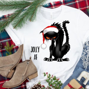 May include: White t-shirt with a black cat wearing a red Santa hat. The cat is looking down with a grumpy expression. The text "Jolly AF" is printed on the shirt.