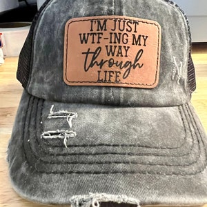Puede incluir: Una gorra de camionero negra desgastada con un parche de cuero marrón que dice "I'm just WTF-ing my way through life".