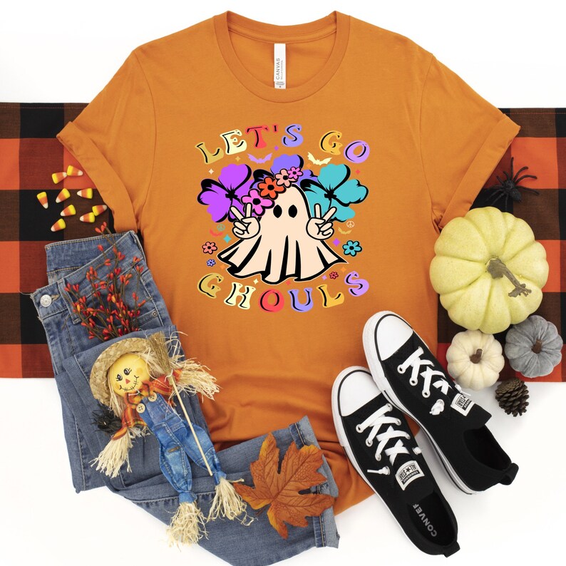 Puede incluir: Una camiseta naranja quemada con un gr&aacute;fico de un fantasma con una corona de flores y signos de paz. El texto "Let's Go Ghouls" est&aacute; impreso en la camiseta.