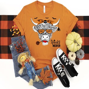 K&ouml;nnte beinhalten: Orangefarbenes T-Shirt mit kurzen &Auml;rmeln und einer schwarz-wei&szlig;en Illustration einer schottischen Hochlandkuh, die eine Sonnenbrille und ein Stirnband mit Flederm&auml;usen und einer Spinne tr&auml;gt. Der Text "Spooky Heifer" ist unter der Kuh gedruckt.