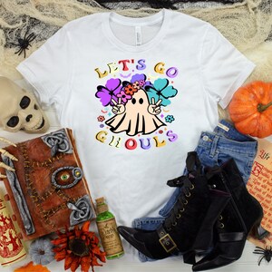 Puede incluir: Camiseta blanca con un gr&aacute;fico colorido de un fantasma con una corona de flores y signos de paz. El texto "Let's Go Ghouls" est&aacute; escrito en letras de colores.