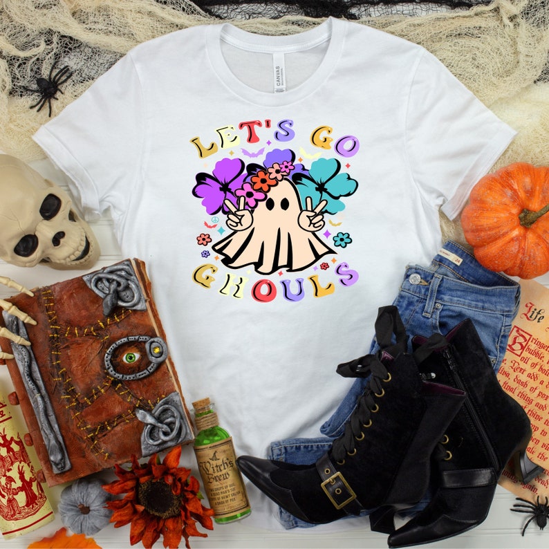 Puede incluir: Camiseta blanca con un gr&aacute;fico colorido de un fantasma con una corona de flores y signos de paz. El texto "Let's Go Ghouls" est&aacute; escrito en letras de colores.