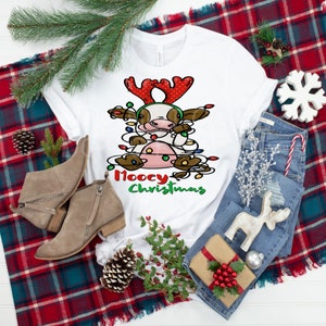 Peut inclure: T-shirt blanc avec une vache de dessin animé portant un bandeau de renne rouge et blanc et emmêlée dans des lumières de Noël. Le texte "Moocy Christmas" est imprimé sous la vache.