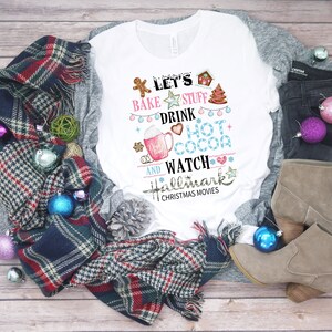 Może przedstawiać: Biała koszula z długim rękawem z świątecznym wzorem, na którym widnieje napis "Let's bake stuff, drink hot cocoa and watch Hallmark Christmas movies". Koszula leży na drewnianej powierzchni z kratkowanym szalem, szyszkami i ozdobami.