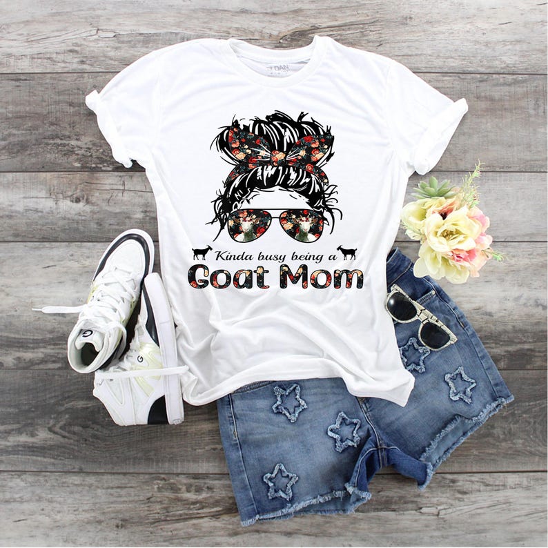 May include: White t-shirt with a floral design of a woman with sunglasses and the text "Kinda busy being a Goat Mom".