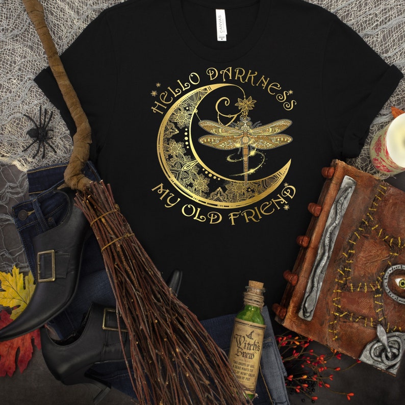 Może przedstawiać: Czarny t-shirt ze złotym wzorem ważki i p&oacute;łksiężyca. Tekst brzmi "Hello Darkness My Old Friend". Koszulka jest częścią kolekcji o tematyce Halloween lub czarownic.