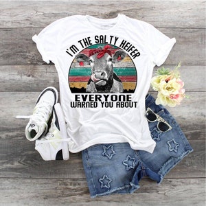 May include: White t-shirt with a retro-style graphic of a cow with a red bandana. The text on the shirt reads "I'm the Salty Heifer Everyone Warned You About".