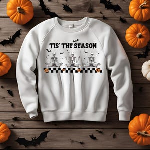 Puede incluir: Sudadera blanca con un gráfico en blanco y negro de tres esqueletos en una pose de yoga. El texto "Tis' the Season" está por encima de los esqueletos. Hay pequeños murciélagos negros y calabazas naranjas en el gráfico.