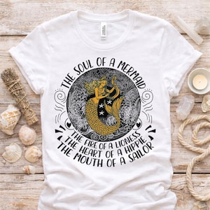 Könnte beinhalten: Ein weißes T-Shirt mit einer schwarz-goldenen Grafik einer Meerjungfrau, die Harfe spielt. Der Text lautet: THE SOUL OF A MERMAID, THE FIRE OF A LIONESS, THE HEART OF A HIPPIE, THE MOUTH OF A SAILOR. Das Design enthält wirbelnde Muster und Sterne.