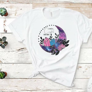 Op de afbeelding: Wit T-shirt met een galaxy-thema halfmaan ontwerp. De maan bevat roze en blauwe kristallen en de tekst "Ik heb een kristal voor dat" is boven de maan gedrukt.