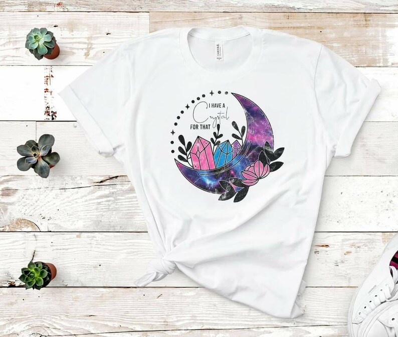 Op de afbeelding: Wit T-shirt met een galaxy-thema halfmaan ontwerp. De maan bevat roze en blauwe kristallen en de tekst "Ik heb een kristal voor dat" is boven de maan gedrukt.