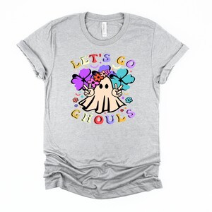 Puede incluir: Una camiseta gris con un fantasma que lleva una corona de flores y signos de paz. El texto "Let's Go Ghouls" est&aacute; impreso en la camiseta.