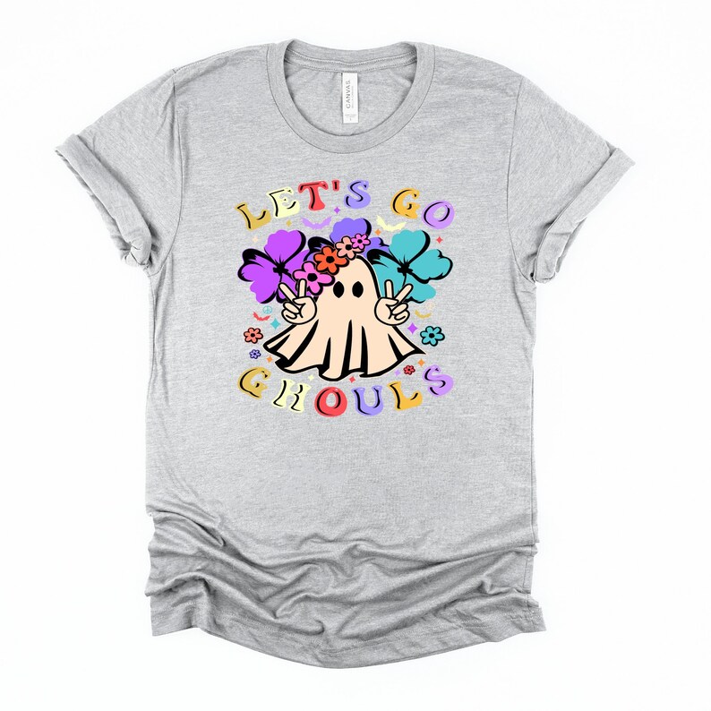 Puede incluir: Una camiseta gris con un fantasma que lleva una corona de flores y signos de paz. El texto "Let's Go Ghouls" est&aacute; impreso en la camiseta.