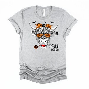 K&ouml;nnte beinhalten: Graues T-Shirt mit einer Schwarzwei&szlig;-Illustration einer Kuh, die eine Sonnenbrille, ein Stirnband mit Totenkopf und gekreuzten Knochen und einen Lolli tr&auml;gt. Der Text "Spooky Heifer" ist unter der Kuh gedruckt.
