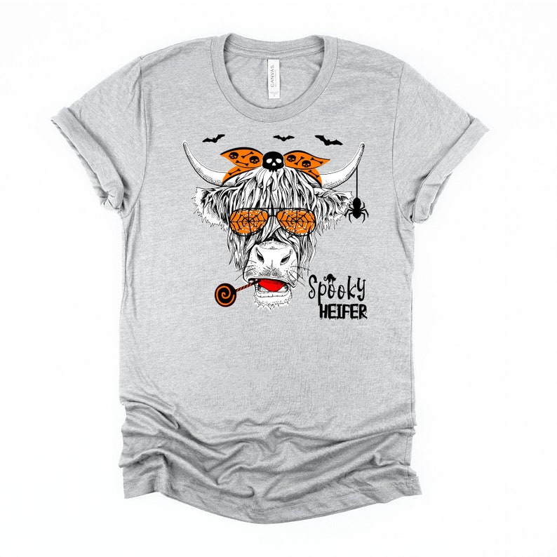 K&ouml;nnte beinhalten: Graues T-Shirt mit einer Schwarzwei&szlig;-Illustration einer Kuh, die eine Sonnenbrille, ein Stirnband mit Totenkopf und gekreuzten Knochen und einen Lolli tr&auml;gt. Der Text "Spooky Heifer" ist unter der Kuh gedruckt.