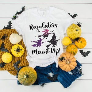 May include: White t-shirt with the text "Regulators Mount Up!" and a graphic of three witches on broomsticks. The shirt is styled with denim shorts, gourds, a mustard-colored knit blanket, and Halloween decorations.