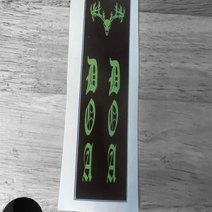 K&ouml;nnte beinhalten: Ein schwarzer und gr&uuml;ner Aufkleber mit einem Hirschsch&auml;del und dem Wort "DOA" in gotischer Schrift.