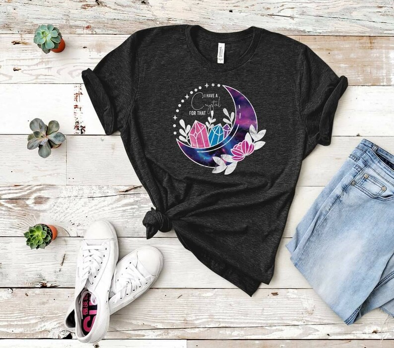Op de afbeelding: Een donker grijs t-shirt met een halve maan-print. De maan is gevuld met een galaxy-print en heeft drie roze en blauwe kristallen erin. De tekst "Ik heb een kristal voor dat" staat op het shirt gedrukt.