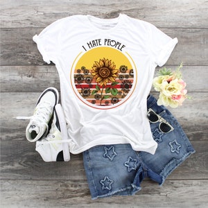 May include: White t-shirt with the text "I HATE PEOPLE" above a sunflower design. The design features a large sunflower in front of a sunset background. The shirt is paired with denim shorts, white sneakers, and sunglasses.