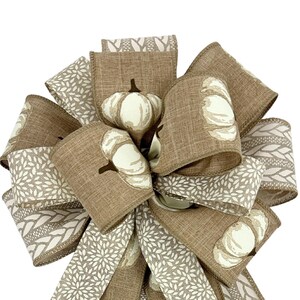 May include: A decorative bow featuring a fall theme. The bow is made of various ribbons in shades of brown and cream. Some ribbons have a pumpkin pattern, while others have floral or woven designs. The bow is suitable for autumn decorations.