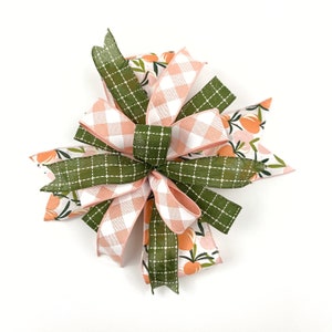 May include: A green and pink bow with a gingham pattern and a peach floral print. The bow is made of ribbon and is tied in a knot.