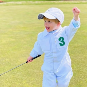 May include: A white jumpsuit with a green number 3 patch on the chest, a matching white baseball cap with a checkered brim, and white shoes. The outfit is worn while holding a black golf club on a green grass field, creating a golf caddy look.