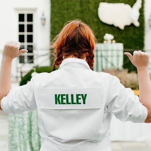 May include: A white long-sleeved jacket featuring a rectangular patch on the back with the name KELLEY printed in bold green capital letters. The jacket has a collared neckline and is worn with matching white pants, viewed from the back.