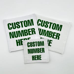 Puede incluir: Tres parches cuadrados de tela blanca con bordes cosidos, cada uno con el texto verde oscuro CUSTOM NUMBER HERE impreso en una fuente negrita sin remates. Los parches están dispuestos en grupo, con dos más grandes y uno más pequeño.