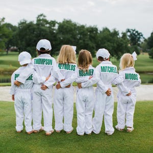 Könnte beinhalten: Sechs Kinder stehen in einer Reihe und tragen passende weiße Langarm-Overalls und weiße Baseballkappen. Jeder Overall hat einen rechteckigen grünen Aufnäher auf der Rückseite, auf dem die Namen Thompson, Reynolds, Rosauer, Mitchell und Woods gedruckt sind.