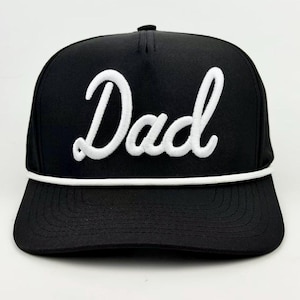 May include: A black baseball cap featuring the word Dad embroidered in white cursive script across the front panel. The cap includes a white cord trim running horizontally across the base of the crown, just above the flat, stitched brim.