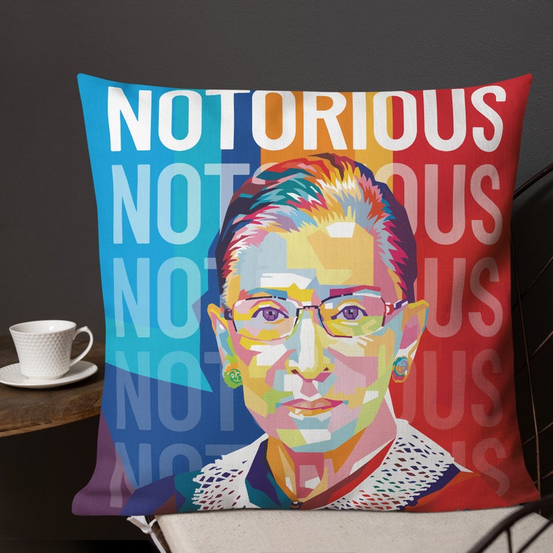 May include: A colorful pop art style pillow featuring a portrait of Ruth Bader Ginsburg with the text "NOTORIOUS" repeated in blue, yellow, and red.