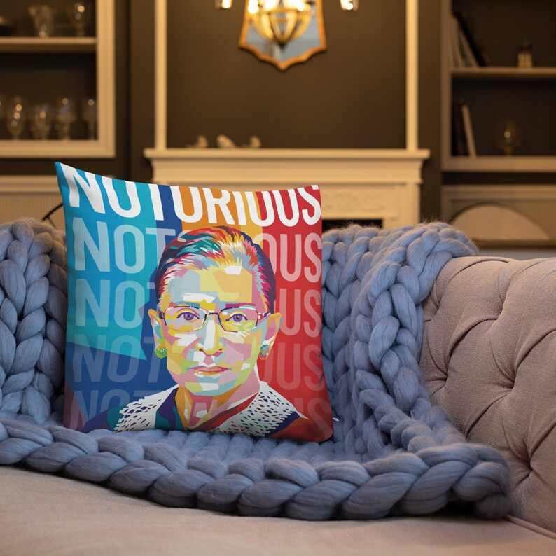 May include: A colorful throw pillow with a portrait of Ruth Bader Ginsburg and the text "Notorious RBG" repeated in a pattern. The pillow is on a blue couch.