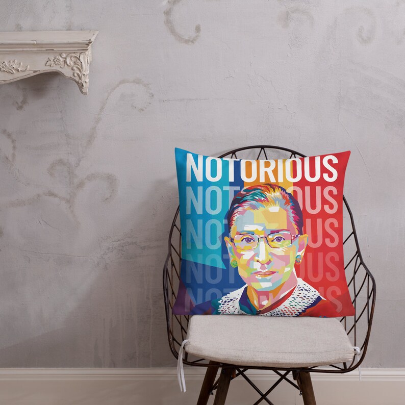 May include: A colorful throw pillow with a portrait of Ruth Bader Ginsburg and the text "NOTORIOUS NOT US" repeated. The pillow is on a white chair with a black metal frame.