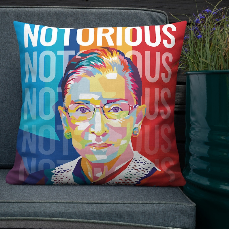 May include: A colorful pop art style pillow featuring a portrait of Ruth Bader Ginsburg with the text "NOTORIOUS" repeated in white on a red, blue, and yellow background.