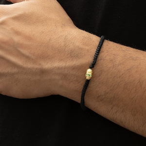 May include: A black cord bracelet with a gold skull charm featuring green gemstone eyes.