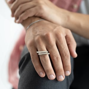 Peut inclure: Une bague en argent avec deux barres parallèles sur la bande. La bague est portée sur le majeur d'une main.