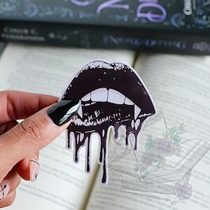 Könnte beinhalten: Ein schwarz-weißer Aufkleber mit Vampirlippen und tropfendem Blut. Der Aufkleber hat scharfe Fangzähne und wird über einem offenen Buch gehalten. Das Design ist im Gothic-Stil gehalten.