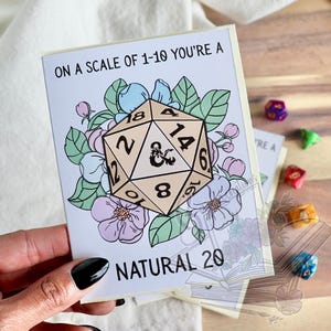 Könnte beinhalten: Grußkarte mit dem Text "ON A SCALE OF 1-10 YOU'RE A NATURAL 20". Die Karte zeigt einen D20-Würfel, umgeben von floralen Illustrationen in Pastellfarben. Zusätzliche Würfel sind im Hintergrund zu sehen.