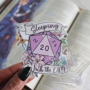 Puede incluir: Una pegatina con un dado morado de veinte caras con el texto "Sleeping With the DM". La pegatina está decorada con elementos florales, una espada y una pancarta. El fondo es un libro abierto borroso.