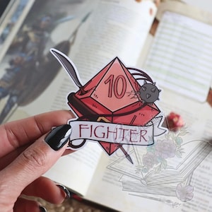 May include: A red and pink sticker featuring a D10 die with the number 10, a sword, and a mace. The word "FIGHTER" is written on a banner below the die. The sticker is held in a hand.