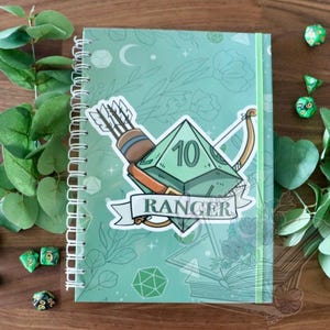Pode incluir: Um caderno de espiral verde claro com uma capa que apresenta um dado D10, um arco e flechas, e a palavra "Ranger". O caderno é decorado com padrões florais e geométricos. Há também vários dados verdes espalhados ao redor do caderno.