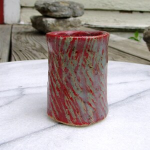 May include: A handmade ceramic tumbler with a cylindrical shape. The tumbler features a vibrant red glaze with swirling streaks of gray and green. The pottery has a rustic, textured appearance, suggesting a handcrafted origin.