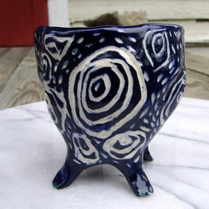 May include: A small, blue and white ceramic pot with three legs and a swirling pattern. The pot is decorated with white swirls on a dark blue background.