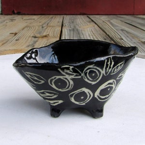 May include: A black ceramic bowl with white floral designs and three legs. The bowl is glazed and has a shiny finish.