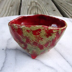 May include: A small, red and green ceramic bowl with an uneven, textured surface. The bowl has a rounded shape and three small feet.