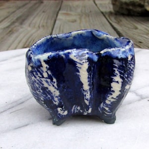 May include: A small, blue and white ceramic bowl with an irregular shape and three legs. The bowl has a textured, hand-painted design.