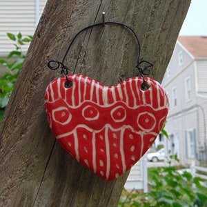 May include: A red and white heart-shaped ceramic ornament with a geometric pattern hangs from a black wire on a wooden surface.