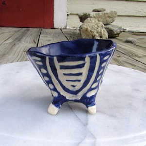 May include: A small, blue and white ceramic bowl with three legs. The bowl has a geometric design and is glazed in a deep blue color.