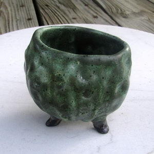 May include: A small, green ceramic pot with a textured surface and three legs. The pot is perfect for planting small plants or succulents.