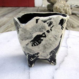May include: A small, black and white ceramic planter with three legs. The planter has a rough, textured surface and is decorated with a white glaze.
